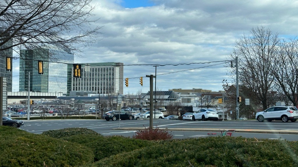 A parking lot full of cars surrounds a shopping mall with skyscrapers in the background