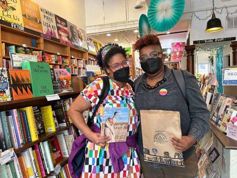 Kristen and Les, masked, standing together holding a book and a Loyalty Bookstore bag inside Loyalty Bookstores Petworth DC location