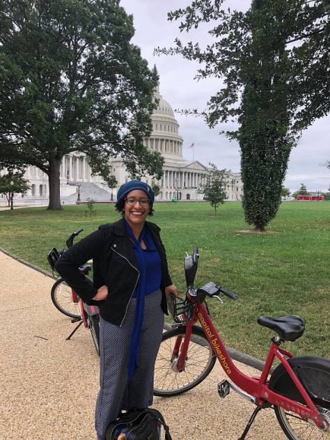 Kristen standing on the East Lawn of the US Capitol, in a blue blouse, a cotton moto jacket and with her hands on her hips, and leaning against a red Captial Bikeshare bicycle. She has on a blue beret and glasses and the US Capitol is in the background on an overcast day.